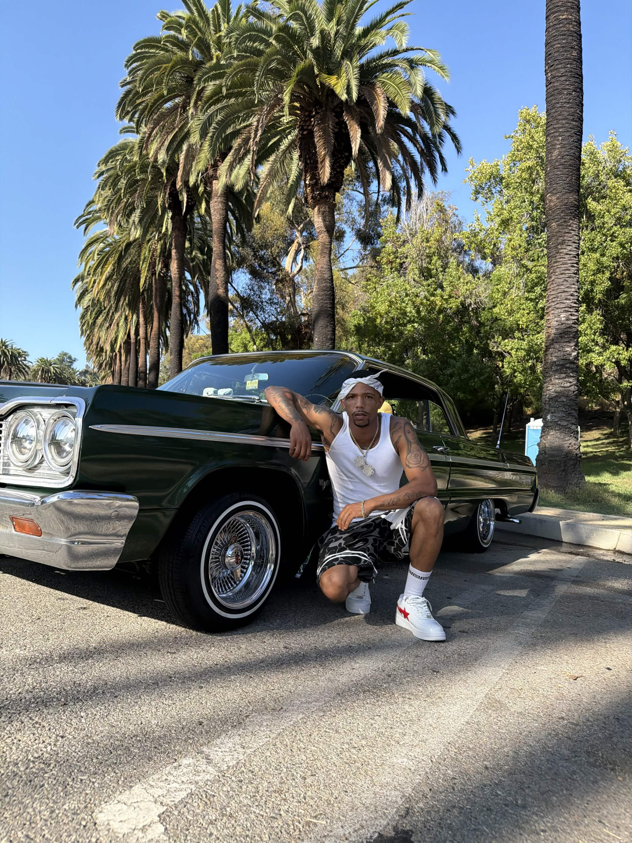 Nhale crouched beside a green lowrider under palm trees